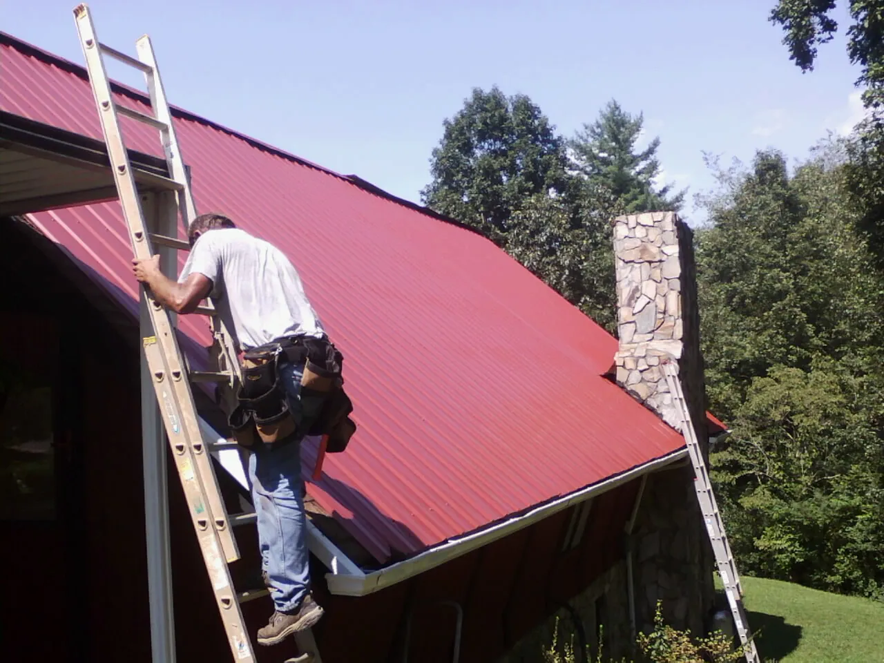 Roofer on a ladder beside a red metal roof and stone chimney — Soffit & Fascia Repair by Roofing Brothers in La Center
